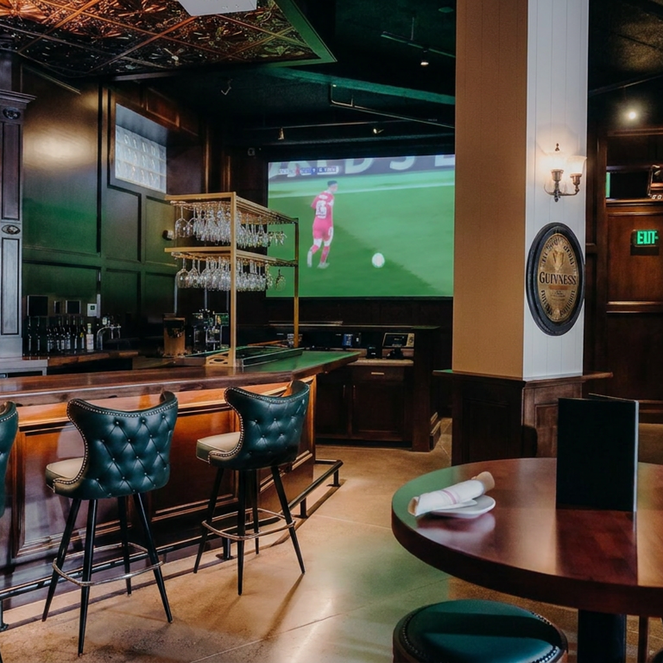 Upscale dark-wood sports bar with leather stools, round dining tables, stocked bar, pool table, and soccer playing on screens.