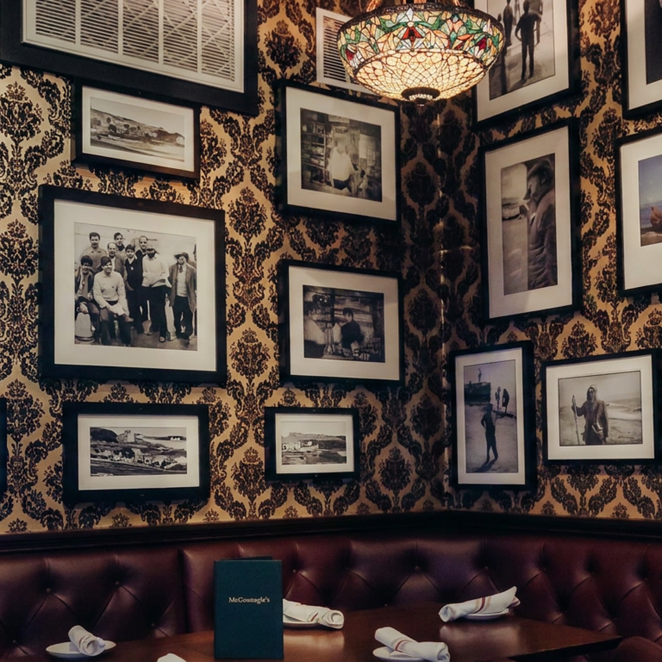 Restaurant dining area with burgundy leather banquettes, patterned wallpaper, framed photographs, stained-glass pendant light, and set tables.