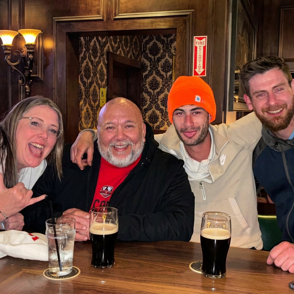 Four smiling friends pose together at a pub table with pints of beer, dark wood paneling, and a sports game on television.