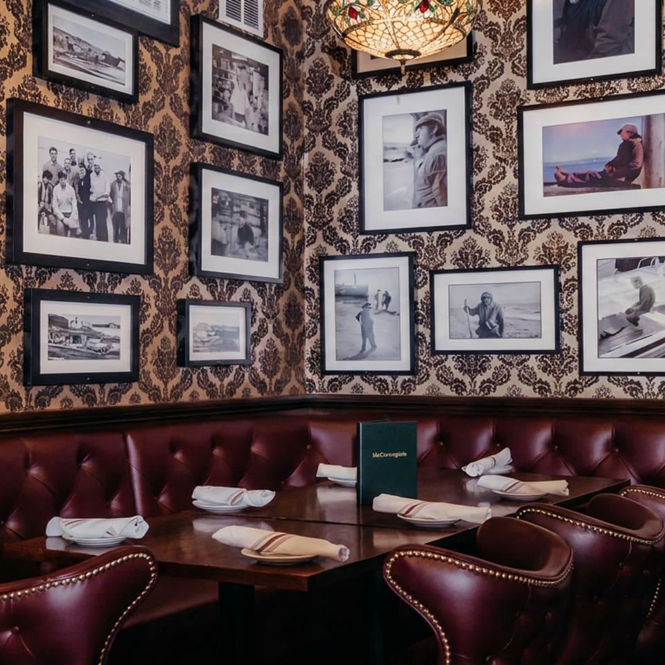 Elegant restaurant dining nook with burgundy leather seating, set tables, ornate wallpaper, framed photographs, and a decorative chandelier.